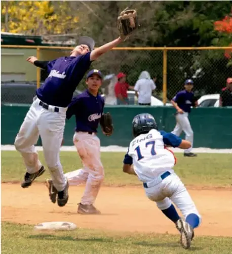 Fotografía de un partido de béisbol: un jugador con uniforme azul salta para atrapar la pelota mientras otro jugador con uniforme blanco y número 17 corre hacia la base.