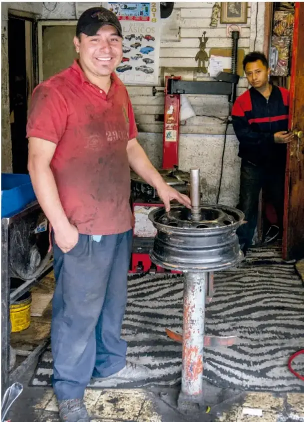 Fotografía de un llantero sonriente con gorra y camiseta roja que sostiene un rin de automóvil en un poste desmontador de llantas; detrás, otro joven en la puerta del taller con llantas y carteles.