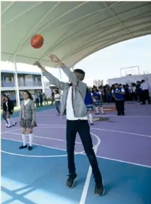 Estudiantes jugando basquetbol en una cancha escolar techada; un joven lanza el balón.