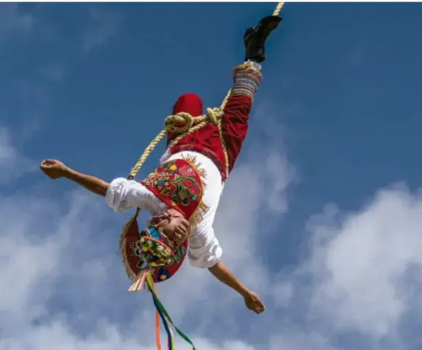 Fotografía de un volador de Papantla colgando invertido de una cuerda durante la danza, con vestimenta tradicional colorida y el cielo de fondo.