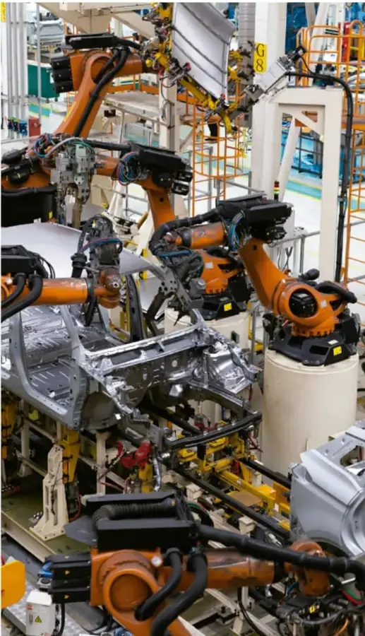 Fotografía de una planta industrial de manufactura automotriz: varios robots industriales naranjas ensamblan carrocerías metálicas de automóviles en una línea de producción automatizada.