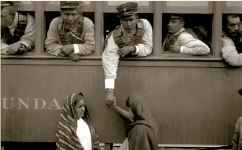 Fotografía histórica en sepia: soldados federales asomados a las ventanillas de un vagón de tren; uno de ellos baja la mano para despedirse de dos mujeres rebozadas que están en el andén. El vagón muestra la palabra 'SEGUNDA'. Pie de foto: 'Soldado federal se despide de mujeres desde la ventanilla de un vagón, 1914. Archivo Casasola.'