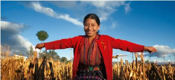 Fotografía de una mujer indígena sonriente con los brazos abiertos en un campo de cultivo, bajo un cielo azul con nubes. Representa la dignidad, la conexión con la tierra y la solidaridad.