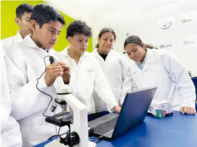 Cinco estudiantes con batas blancas de laboratorio observan al microscopio y trabajan con una computadora portátil sobre una mesa azul.