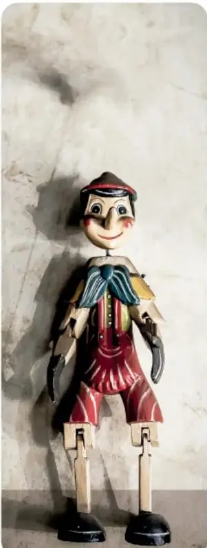 Fotografía de un muñeco de madera articulado con sombrero, chaleco y pantalones rojos, apoyado sobre una superficie de mármol, proyectando una sombra en la pared.