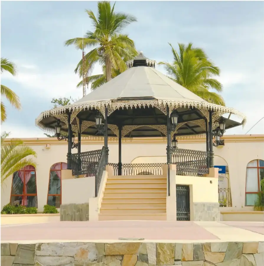 Fotografía del kiosco de Todos Santos: estructura octagonal de hierro forjado con doble techo, faroles colgantes y escalinata central, rodeado de palmeras altas y edificios claros con arcos, bajo un cielo azul.