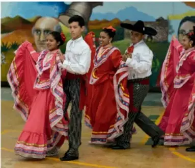 Fotografía de un grupo de jóvenes bailando un tradicional baile folclórico con vestidos largos rojos y blancos, sombreros y faldones bordados, sobre un escenario; representa las producciones artísticas y la cultura comunitaria de la que se nutre el proyecto.