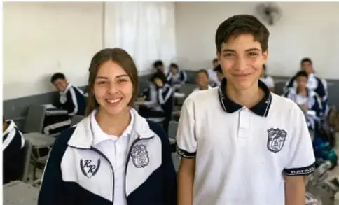 Fotografía de dos estudiantes de secundaria de pie en un salón de clases, con compañeros sentados al fondo; crédito a Perla Arell Zúñiga Aguilar, ZALATIOS.