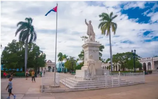 Fotografía de una plaza con una estatua de José Martí, una bandera cubana y palmeras, en Cuba.