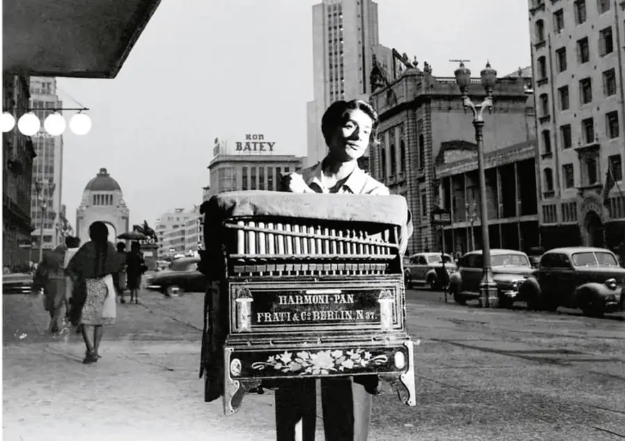 Fotografía en blanco y negro, aparentemente antigua, de un organillero que carga su organillo marca 'Harmoni-Pan, Frati & Co. Berlin N.7' en una avenida con edificios altos, autos antiguos y peatones; al fondo se ve un monumento.