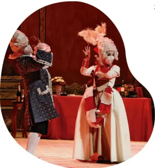 Fotografía de una representación teatral con dos actores enmascarados con pelucas y vestimenta barroca (una con abanico, otra con traje rojo y blanco) frente a un mantel rojo, en un escenario iluminado.