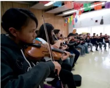 Fotografía de un grupo de estudiantes en un salón multicolor; una niña en primer plano toca el violín mientras el resto del grupo participa en la actividad musical.