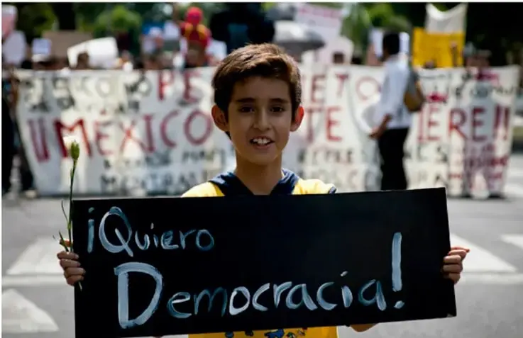 Fotografía de un niño sosteniendo un cartel que dice '¡Quiero Democracia!' con una flor en la mano, en medio de una marcha con pancartas de fondo.