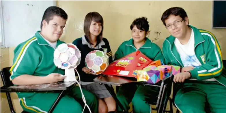 Fotografía de cuatro estudiantes con uniforme verde mostrando sus productos del proyecto: esferas decoradas tipo maqueta y una maqueta de edificio, sentados en su aula.