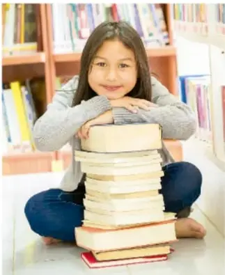 Niña sonriente sentada junto a una pila de libros en una biblioteca.