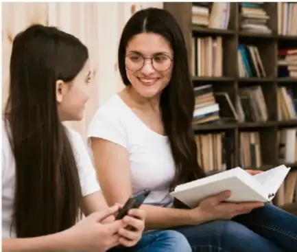 Fotografía de una docente y una estudiante conversando con un libro, en una biblioteca con estanterías de libros al fondo.