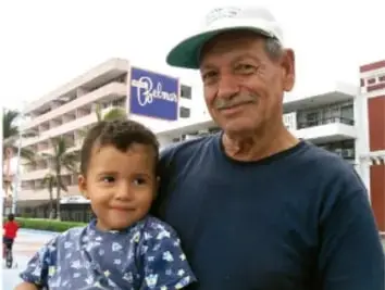 Fotografía de un adulto mayor con gorra y un niño pequeño posando frente a un edificio con el letrero 'Prisma'; alude a los narradores o comunidades que comparten anécdotas.
