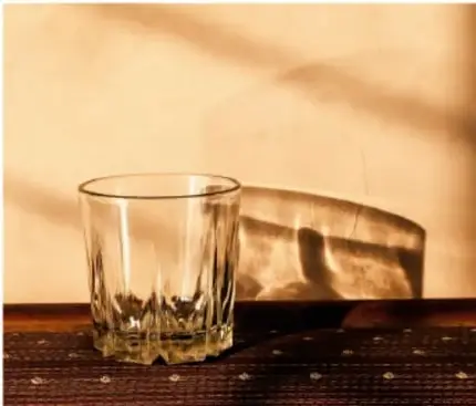 Fotografía de un vaso de vidrio transparente junto a un recipiente más opaco, con rayos de luz del sol atravesándolos sobre una mesa de madera.