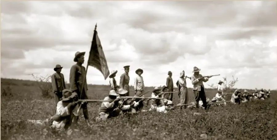 Fotografía histórica en sepia de tropas constitucionalistas durante la batalla de Celaya, abril de 1915: soldados en campo abierto, algunos en posición de tiro con fusiles, uno porta una bandera; el cielo está nublado. Crédito: (5718) Secretaría de Cultura, INAH, SINAFO.FN.MX.