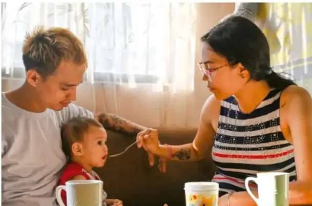 Fotografía de una familia (un joven, una mujer y una niña pequeña) en casa, dando de comer a la niña, mientras conversan.
