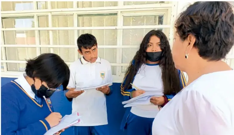 Fotografía de tres estudiantes de telesecundaria con uniforme y cubrebocas entrevistando a una mujer adulta en el patio de su escuela, tomando notas en cuadernos. Crédito: Luis Eduardo García Medel, Tlaxcala.