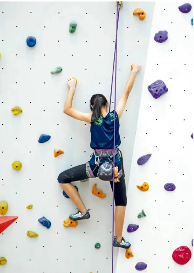 Fotografía de una mujer escalando un muro de escalada con agarres de colores y cuerda de seguridad; representa avanzar poco a poco ante un reto.