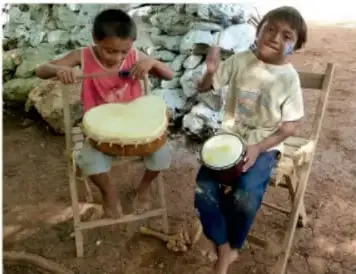 Dos niños sentados tocan tambores artesanales; uno viste playera rosa y el otro playera clara, en un entorno rural.