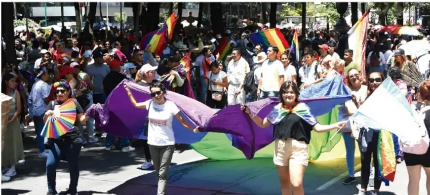 Fotografía de una marcha pacífica en la que jóvenes y adultos caminan sosteniendo una gran bandera con los colores del arcoíris, en un entorno urbano arbolado. La imagen ilustra la participación juvenil en espacios públicos y la diversidad.