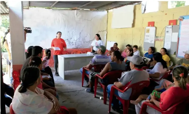 Fotografía de un grupo de personas reunidas en un espacio techado comunitario, sentadas en sillas rojas frente a dos personas que conducen la sesión; representa una actividad de recogida de información o asamblea vecinal.