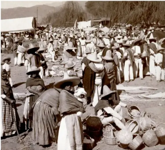 Fotografía histórica en blanco y negro que muestra a un grupo numeroso de personas, en su mayoría mujeres con vestimenta tradicional y sombreros, reunidas en un mercado o plaza al aire libre. Se observan canastas y puestos de venta, lo que refleja la vida cotidiana y el comercio popular de la época.