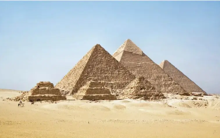 Dos pirámides nubias delgadas en el desierto de Sudán: una completa de talud empinado y otra deteriorada; junto a ellas, pequeñas construcciones con puertas decoradas.