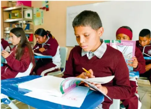 Fotografía de estudiantes de telesecundaria con uniforme rojo trabajando en el aula, leyendo libros y escribiendo en sus cuadernos.