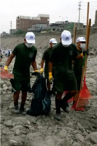 Fotografía de un grupo de estudiantes con camiseta verde, gorra blanca, guantes y cubrebocas recogiendo residuos con bolsas y rastrillos en un terreno pedregoso junto a un cauce o río, durante una jornada de limpieza ambiental escolar.