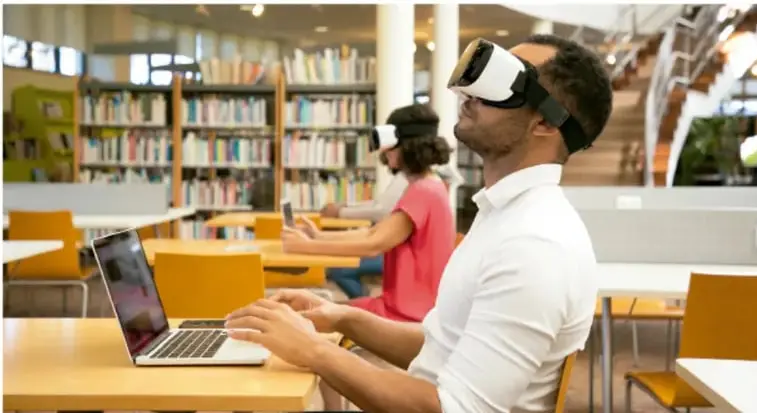 Fotografía de usuarios en una biblioteca o espacio de estudio: una persona en primer plano usa lentes de realidad virtual frente a una computadora portátil, mientras otra persona al fondo también usa un visor de realidad virtual.