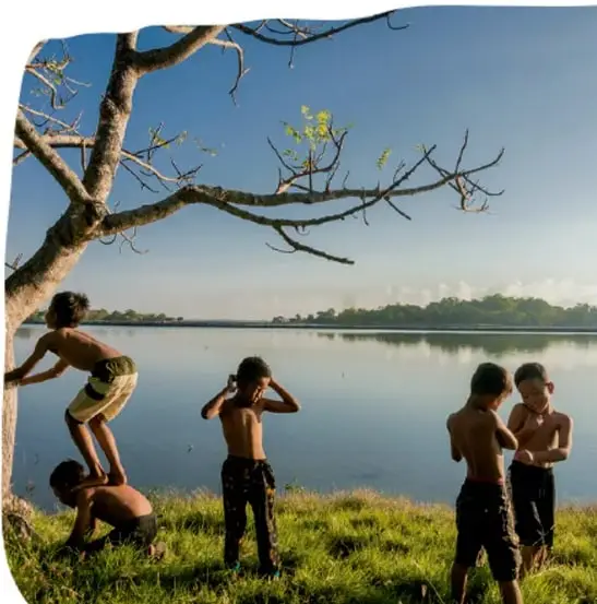 Fotografía de niños jugando a orillas de un río o lago, con un árbol en primer plano; dos de ellos forman una pirámide humana y otros se observan, ilustrando el juego y la actividad motriz al aire libre.
