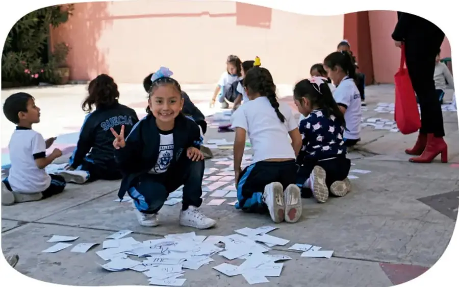 Fotografía de niñas y niños de primer grado sentados en el patio de la escuela, jugando con tarjetas de papel sobre el suelo; una niña sonríe y hace el gesto de paz con la mano, y una adulta con bolsa roja los acompaña.