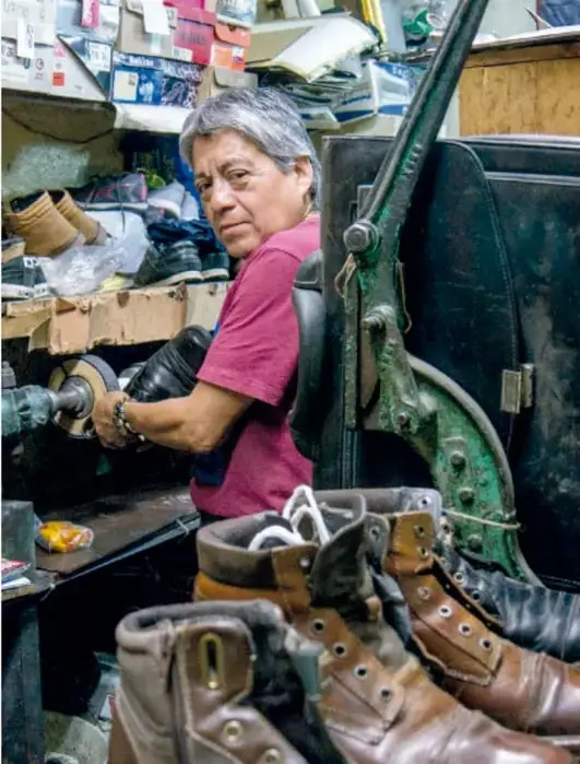 Fotografía de un maestro zapatero en su taller, frente a una máquina antigua de coser cuero verde, rodeado de botas y zapatos de piel, con cajas y herramientas en las paredes.