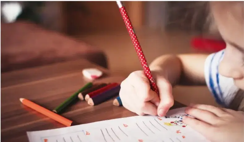 Fotografía de una niña escribiendo con un lápiz rojo sobre una hoja con renglones; a su lado hay lápices de colores sobre una mesa de madera.