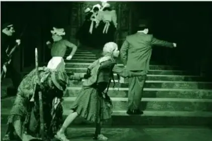 Fotografía teatral a color, en tonos oscuros: escalera en una acera que sube hacia puertas de un recinto; ocho personajes enmascarados, entre ellos una niña con trenzas, una persona con harapos y báculo, un señor con portafolio, un policía, una empleada doméstica y tres mucamas; una luz ilumina la escena.