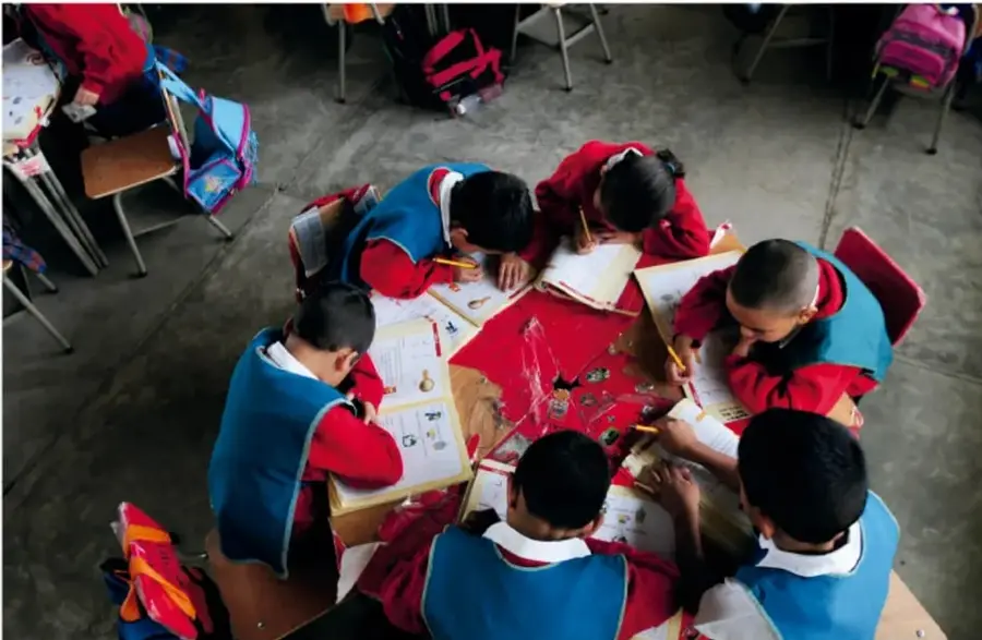 Estudiantes de primaria con uniforme escolar sentados en círculo alrededor de mesas, trabajando en equipo con cuadernos y materiales.