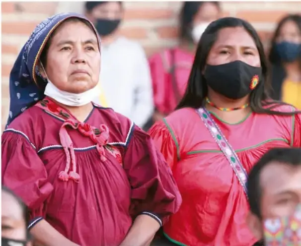 Fotografía de mujeres indígenas mexicanas con trajes tradicionales coloridos; una de ellas usa mascarilla, otra lleva rebozo y cabello trenzado con cinta, en un acto público o comunitario.