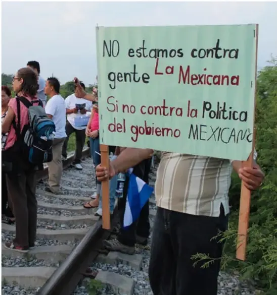 Fotografía de una persona sosteniendo un cartel en una protesta. El cartel dice: 'NO estamos contra gente La Mexicana Si no contra la Política del gobierno MEXICANO'. Se observan vías de tren y otras personas alrededor.