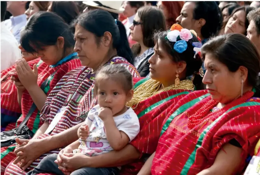 Fotografía en color de mujeres indígenas de Guatemala o México, vestidas con huipiles rojos tradicionales, sentadas en fila participando en una asamblea o reunión comunitaria; una de ellas sostiene a un niño pequeño. Ilustra la participación comunitaria.