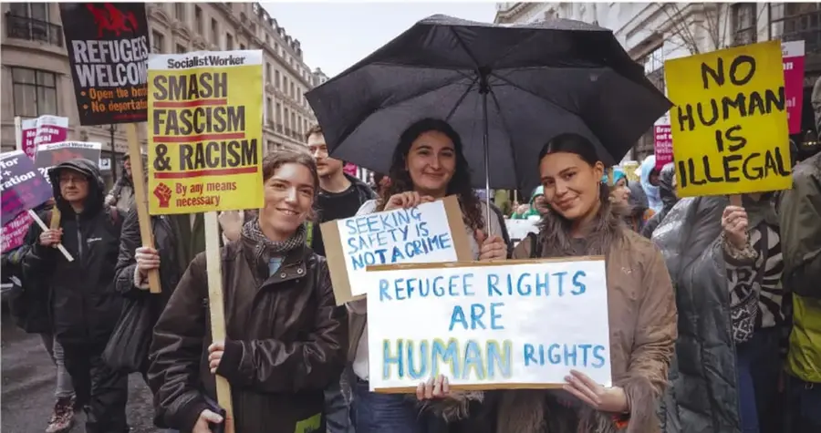 Fotografía de una marcha actual por los derechos humanos: varias personas, algunas con paraguas, sostienen pancartas con lemas en inglés como 'Refugee rights are human rights', 'No human is illegal', 'Smash fascism & racism' y 'Seeking safety is not a crime'.
