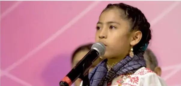 Fotografía de una niña hablando por un micrófono durante una asamblea, con otras personas al fondo.