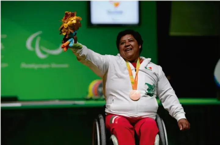 Fotografía de una mujer deportista en silla de ruedas, con medalla y ramo de flores, celebrando en un evento deportivo internacional. Representa la inclusión y la participación de personas con discapacidad.