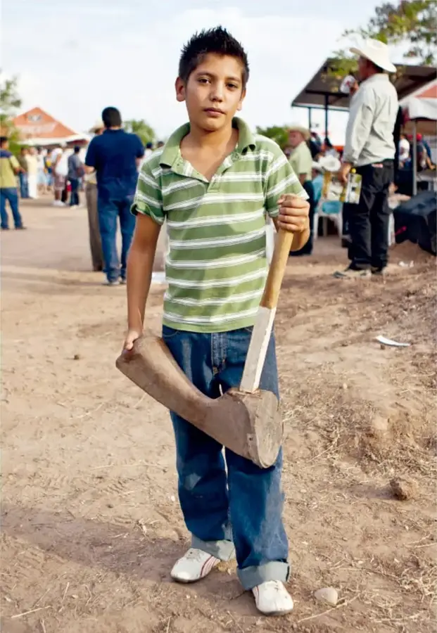 Niño con playera verde a rayas y pantalón de mezclilla sosteniendo un mazo de madera en un espacio abierto de tierra; al fondo, personas y puestos.