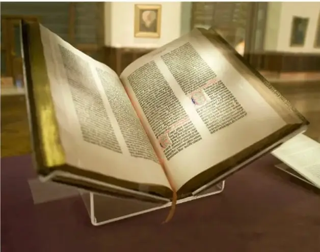 Fotografía de un libro antiguo abierto sobre un atril de exhibición en una vitrina, con páginas de doble columna de texto impreso en tinta oscura y una inicial ornamentada en rojo; al fondo se aprecia el interior de una biblioteca o sala de consulta con estanterías y un retrato enmarcado en la pared.