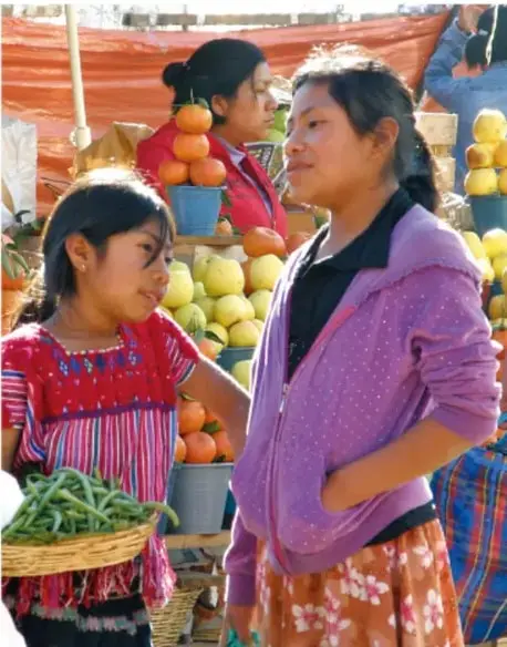 Fotografía de dos mujeres en un mercado: una joven con blusa bordada sostiene una canasta con verduras y una mujer adulta con chamarra morada observa; al fondo hay frutas y otra persona. Representa el trabajo cotidiano y la diversidad cultural.
