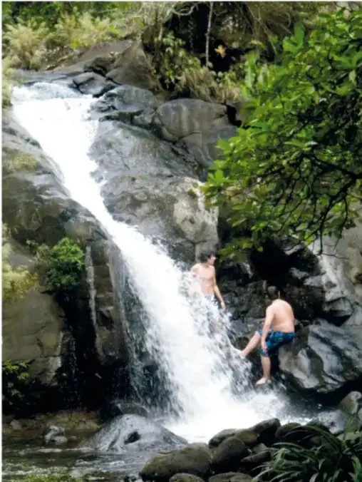 Cascada entre rocas cubiertas de vegetación; dos personas jóvenes se asoman entre las piedras del chorro de agua.